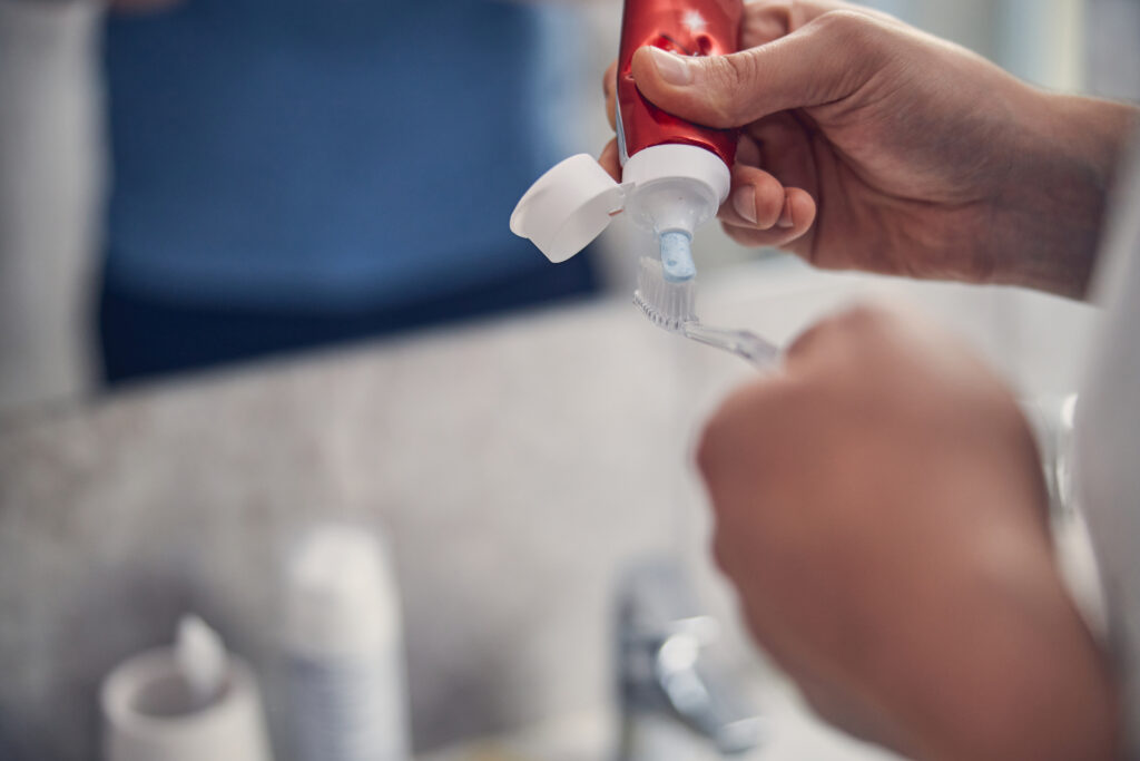 Close-up of a hand squeezing fluoride toothpaste onto a toothbrush over a bathroom sink