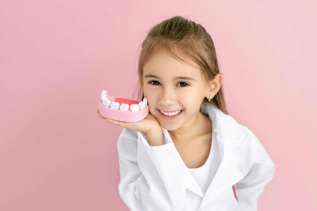 Smiling young girl in a white coat holding a pink model jaw beside her face, illustrating fissure sealants for children’s teeth